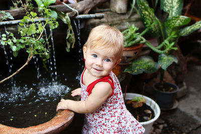 Little smiling girl with blond hair in sundress is standing in garden with pot plants and waterfall.