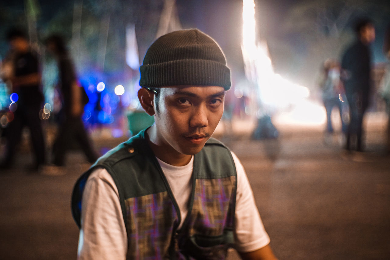 portrait, looking at camera, incidental people, focus on foreground, lifestyles, young adult, real people, leisure activity, night, front view, one person, hat, headshot, illuminated, young men, city, casual clothing, clothing, street