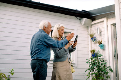 Senior man talking with woman holding mobile phone against house