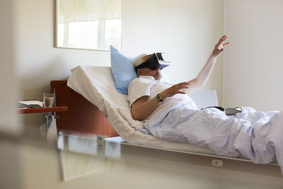 Senior man gesturing while using vr glasses on bed in hospital ward