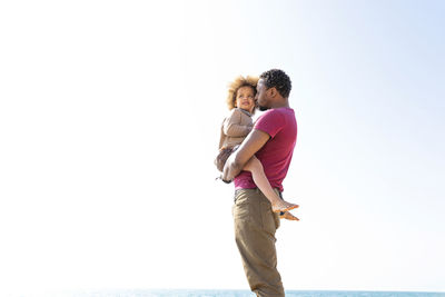 Father carrying daughter standing in front of sky on sunny day
