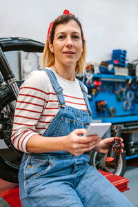Mechanic woman looking phone and holding beer sitting in front of motorcycle on factory