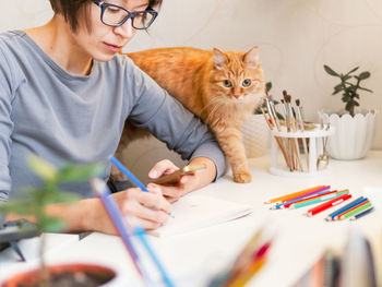 Woman with short hair cut is drawing in notebook. ginger cat sits near her. fluffy pet and artist. 