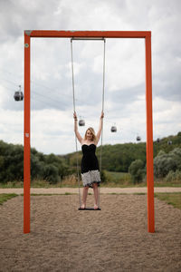 Full length portrait of woman standing on swing against sky