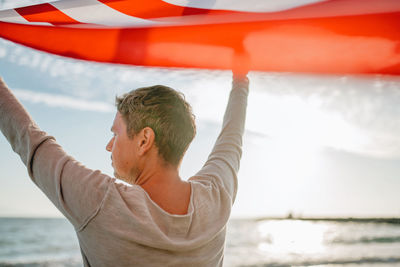 Rear view of man holding american flag while looking at sea