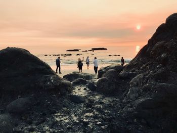 People on rocks at beach against sky during sunset