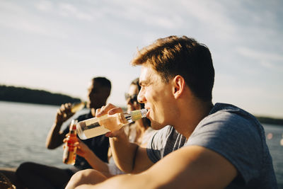 Man enjoying drink with friends while sitting on jetty against sky in summer