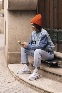 Full body of smiling female in casual wear and hat text messaging on smartphone while sitting on skateboard near building in city