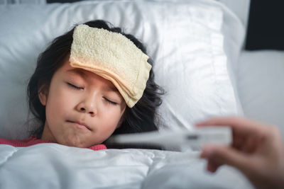 Cropped hand holding thermometer while girl resting on bed