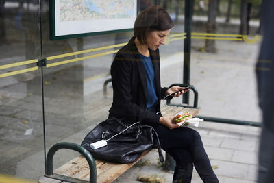 Mid adult businesswoman using smart phone while eating sandwich at bus stop in city