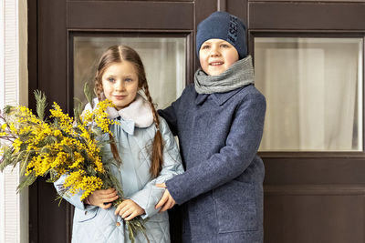 A couple of a boy and a girl of schoolchildren in spring coats with a bouquet of mimosa in  hands 