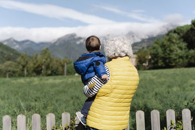 Rear view of a grandmother wearing a medical mask carrying her little grandson in a field