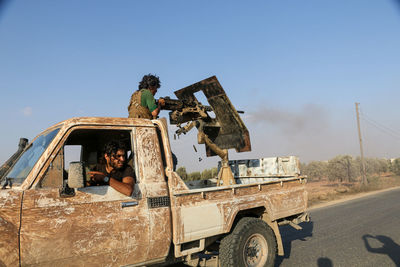 Soldiers in pick-up truck on road against clear sky