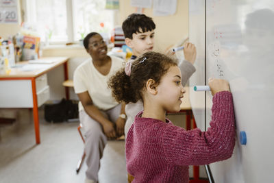 Girl and boy writing on whiteboard while solving mathematics in classroom