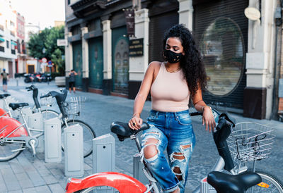 Side view of charming young latin female in jeans and protective mask standing near bicycle on city street and looking away