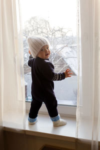 Child boy in a warm blue jumpsuit is on window in winter white knit hat