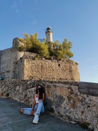 Full length of woman sitting on stone wall by building against sky