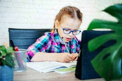 Little girl in eyeglasses making notes in her noteblook whlile studying online at home