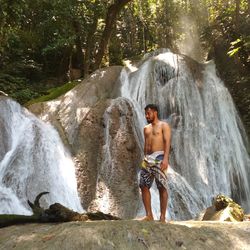 Full length of shirtless man standing against waterfall in forest