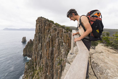 Man carrying a backpack leans over the top of sea cliff in tasmania.