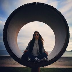 Portrait of young woman sitting in concrete pipe at beach against sky