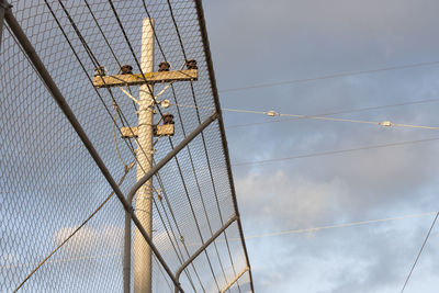 Low angle view of chainlink fence by telephone pole against cloudy sky
