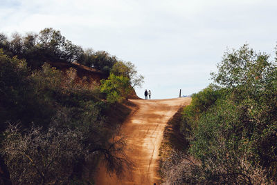 People walking on dirt road amidst trees against sky