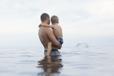 Side view of shirtless father carrying son while standing in sea against cloudy sky