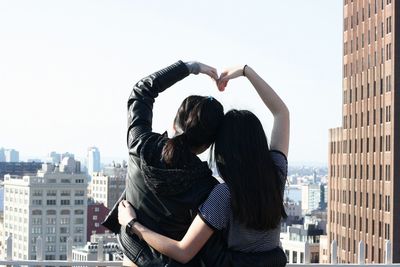 Rear view of friends making heart shape in city against clear sky