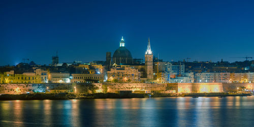 Illuminated buildings by river against blue sky