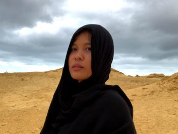 Portrait of young woman in hijab standing on land against cloudy sky