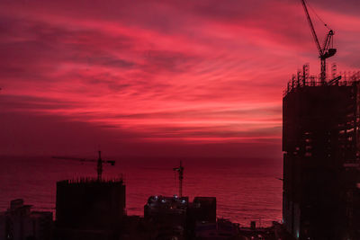 Silhouette buildings by sea against dramatic sky during sunset