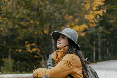 Side view portrait of young woman leaning on wooden fence by road in autumn woods