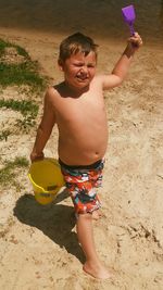 Full length of shirtless boy holding sand pail and shovel while walking at beach during summer