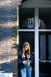 Concentrated young ethnic female traveler with long dark hair in casual outfit leaning on brick wall of aged building and watching photos on digital camera on sunny day in amsterdam