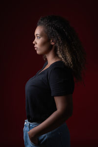 Side view of young overweight african american woman in casual black shirt and jeans standing in red studio looking away