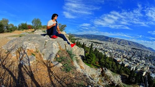 Man looking at cityscape while sitting on rock formation against sky