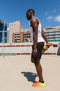 Side view of fit black male athlete stretching legs while warming up muscles before workout on sports ground on sunny day