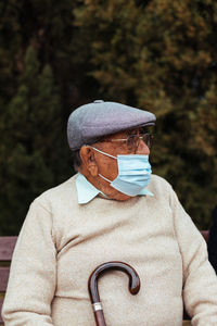 Portrait of elderly man with face mask and beret looking away while sitting on a park bench