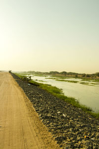 Scenic view of dirt road by lake against sky