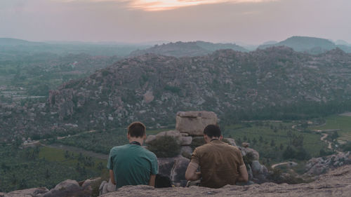 Rear view of men sitting on rock against sky