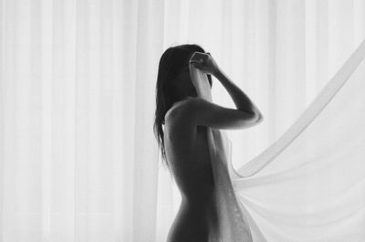 Side view of naked young woman standing by curtain at home