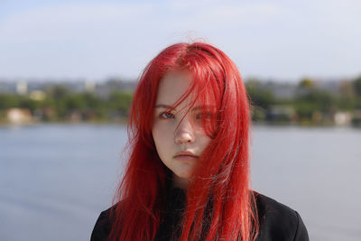 A young girl with a pierced nose and red hair stands on  the background of a river on a sunny day