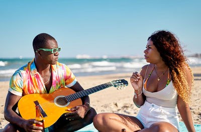 African american male musician in stylish colorful shirt and sunglasses drinking beer while sitting on sandy beach with acoustic guitar and talking to girlfriend during summer vacation