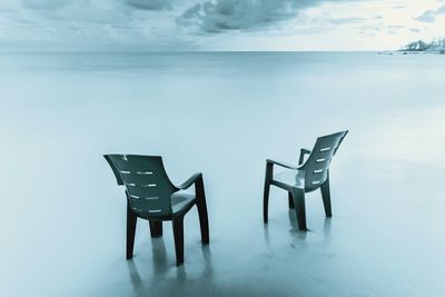 Empty chairs and table by sea against sky