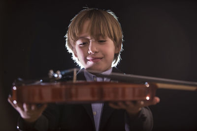 Boy holding violin while sitting against black background