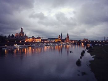 Illuminated buildings by river against cloudy sky 