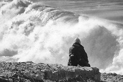 Rear view of person sitting on rock in front of wave at sea shore