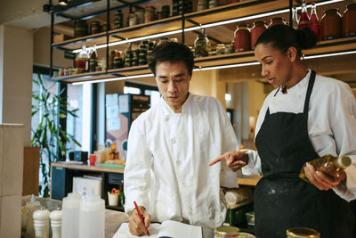 Male chef taking inventory with female chef holding bottle while standing in commercial kitchen