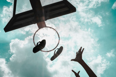 Low angle view of cropped hands by basketball hoop and flip-flops against sky